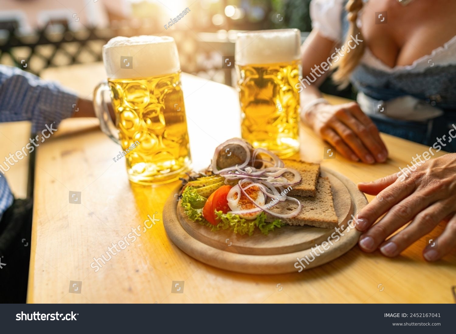 Traditional Bavarian Obatzda with pretzels and radishes and beer mugs, man and young woman in tracht in the background at beer garden or oktoberfest, Munich, Germany #2452167041