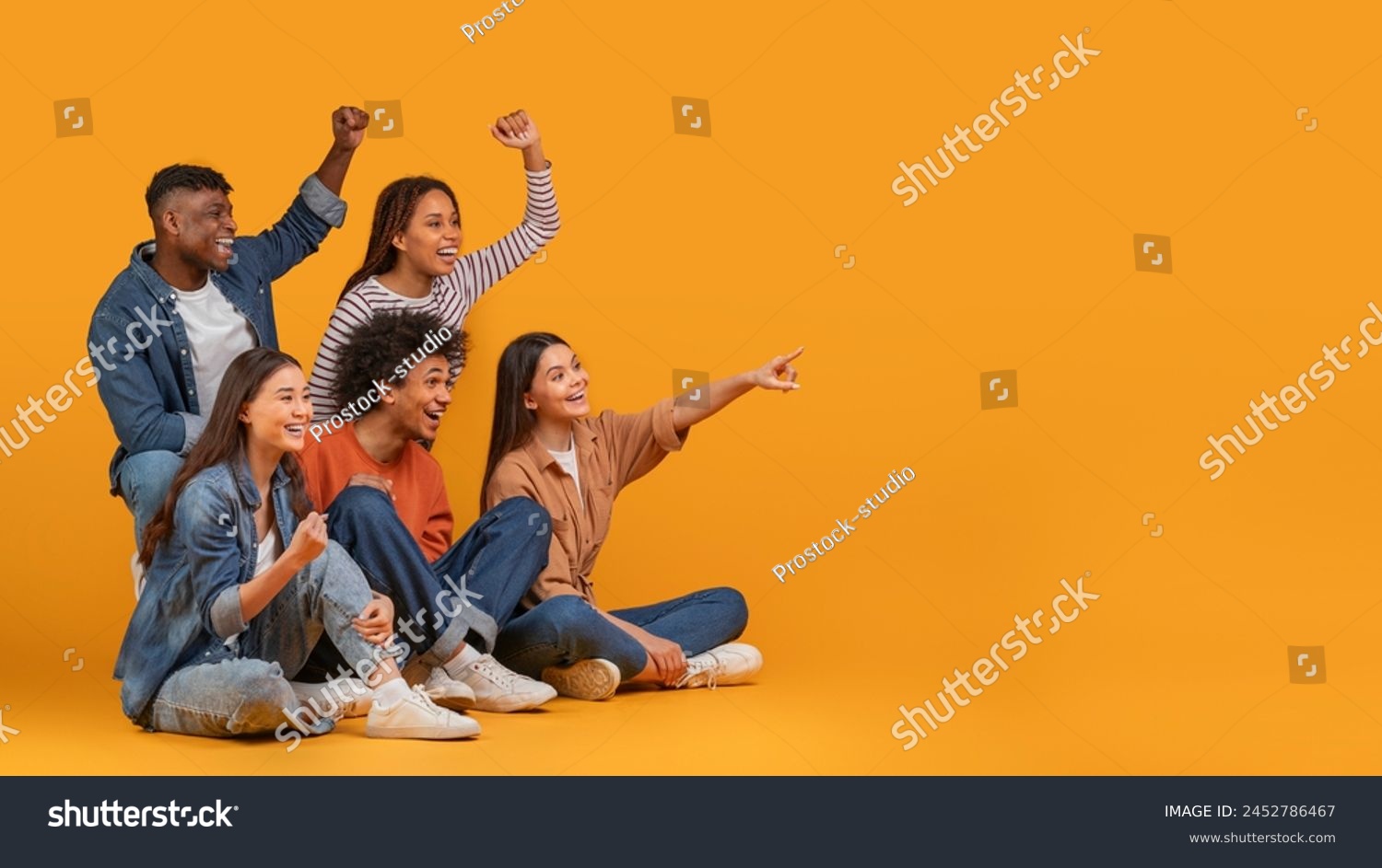 Multiethnic friends excitedly cheering and pointing to the side, exemplifying celebration and happiness in a diverse group, isolated on a yellow background, copy space #2452786467