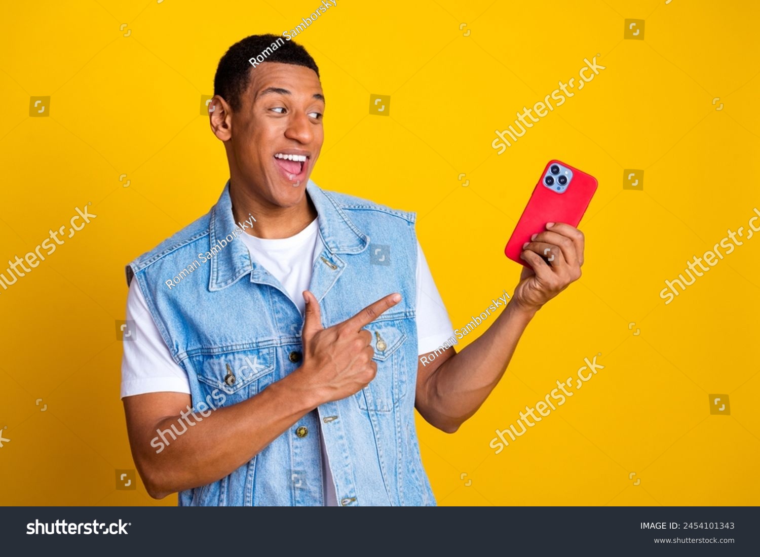 Portrait of multiethnic multinational man dressed denim vest over t-shirt directing look at smartphone isolated on yellow color background #2454101343