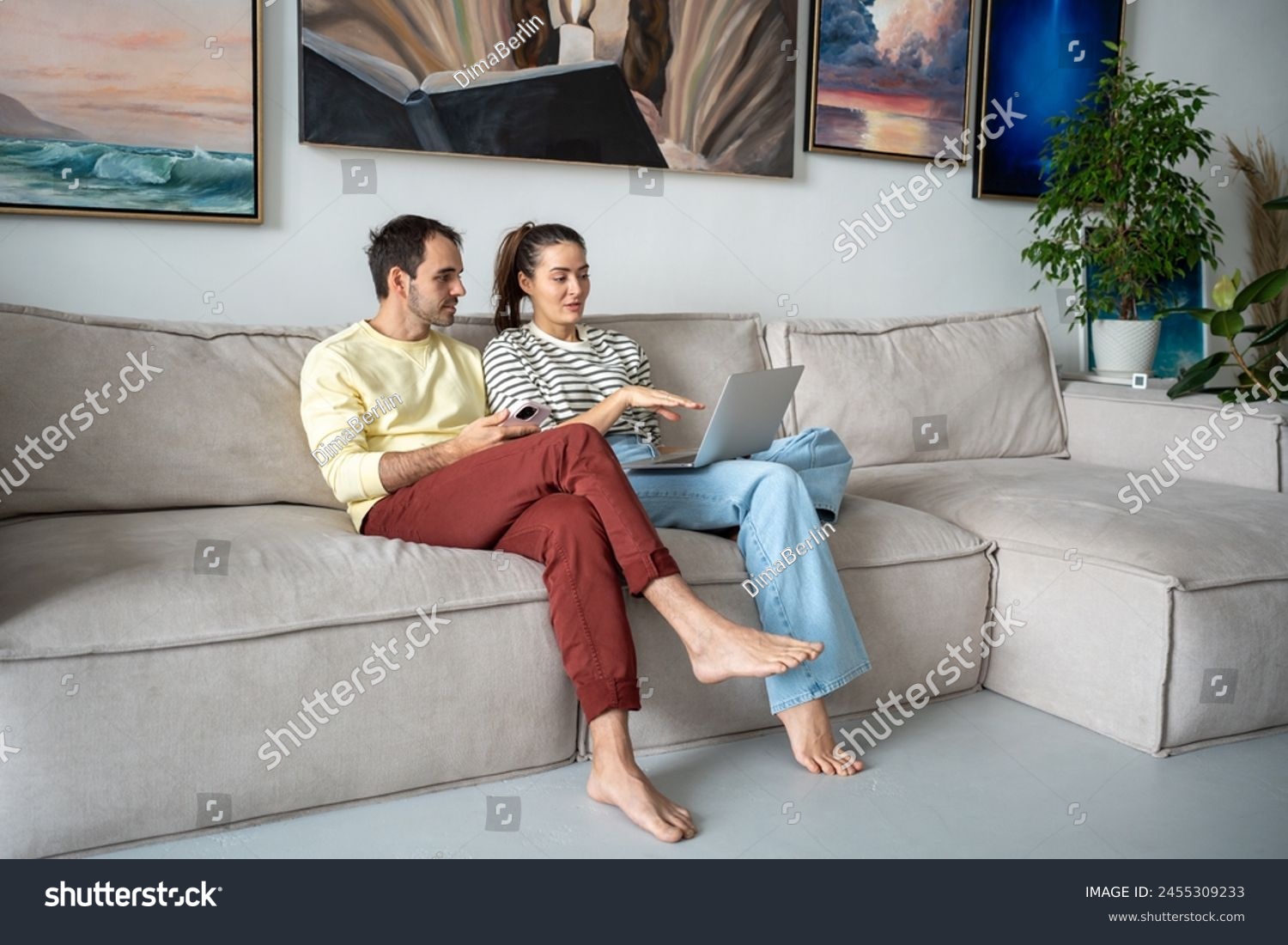 Pleasant family couple artists sits on couch looking at laptop screen browsing internet together. Creative young married spouse web surfing, making purchases online, booking flight tickets for travel. #2455309233