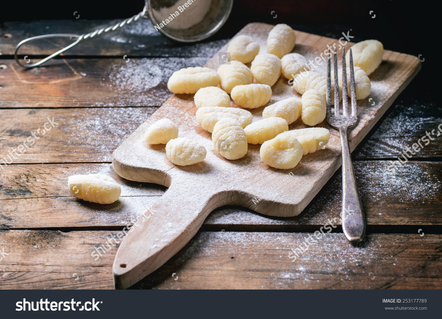 Uncooked homemade potato gnocchi with fork and strainer on vintage cutting board over wooden table with flour. See series. #253177789