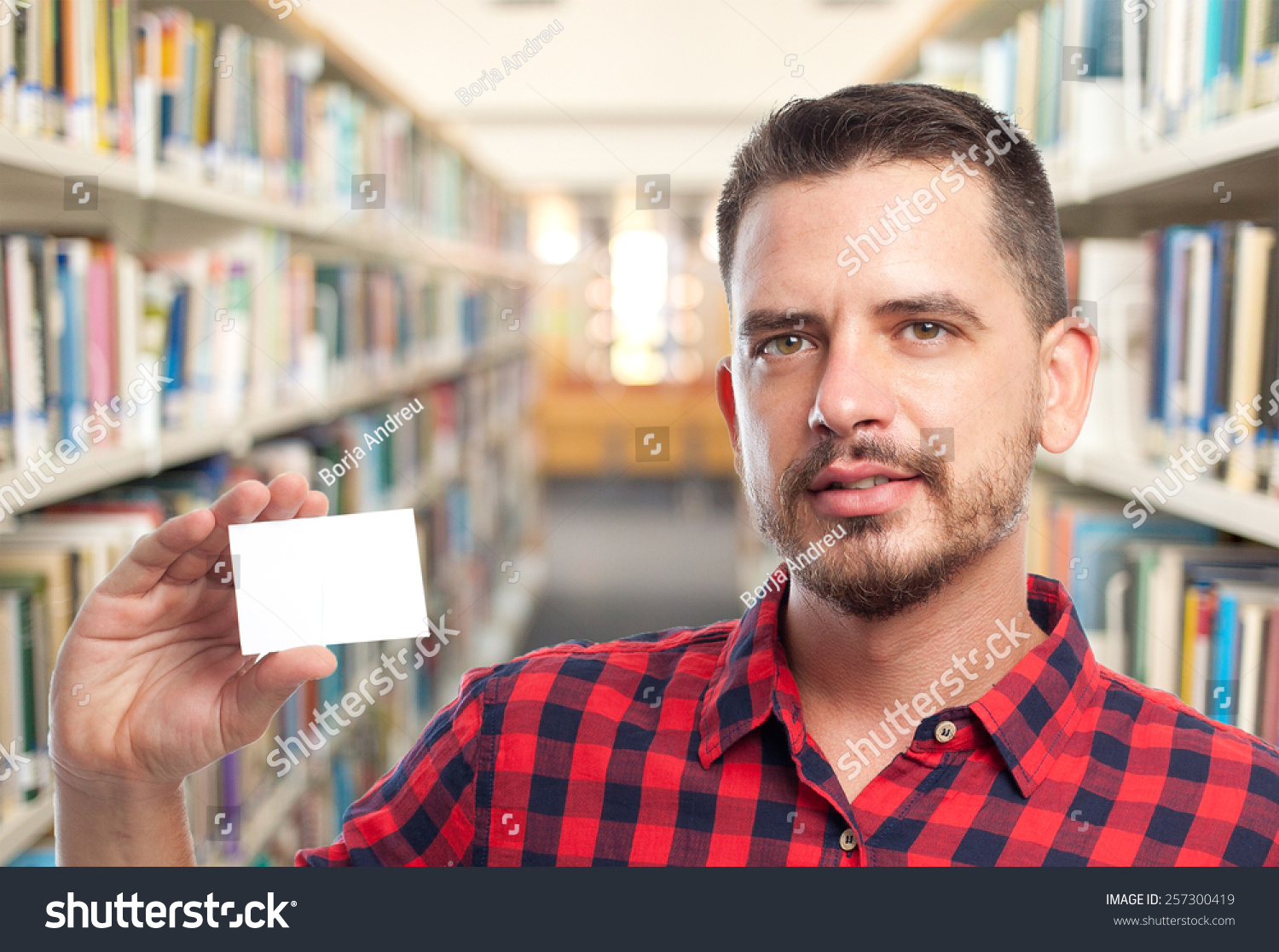 Man with red squares shirt. He is holding a white card. Over library background #257300419