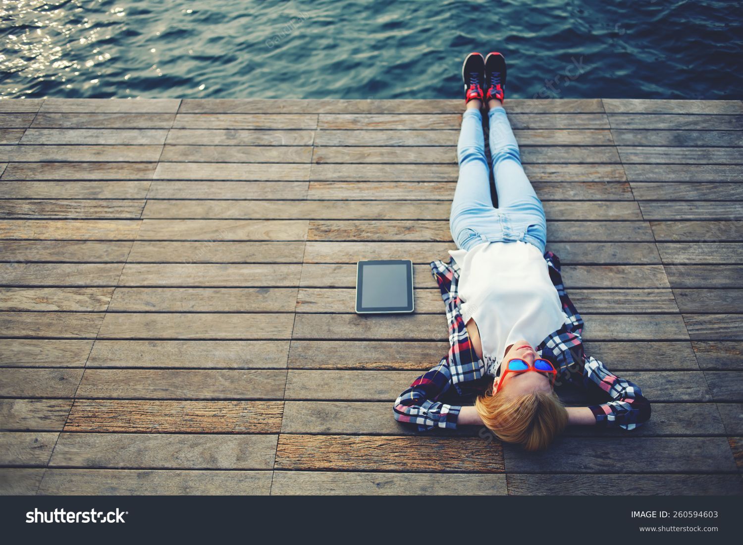 Top view young woman lying on a wooden jetty enjoying the sunshine,tourist girl in bright glasses lying on jetty by river, vintage photo of relaxing young woman in nature with tablet, cross process #260594603