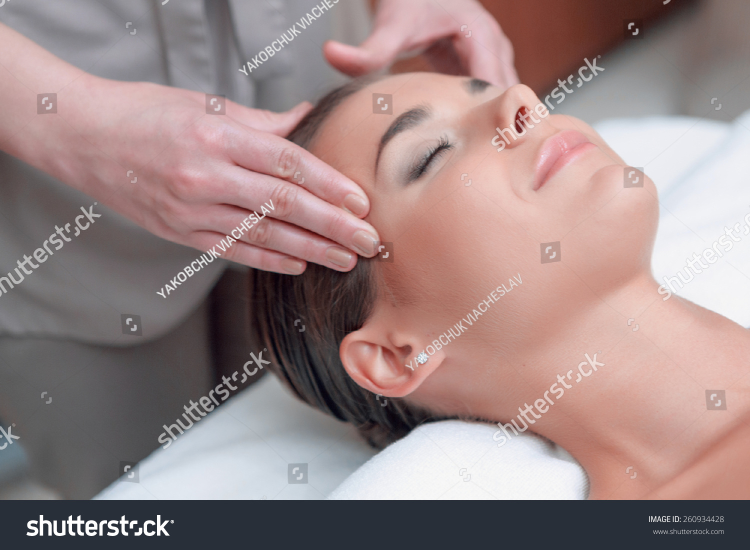 Facial massage. Top view of attractive young woman lying on back and keeping her eyes closed while massage therapist massaging her face #260934428