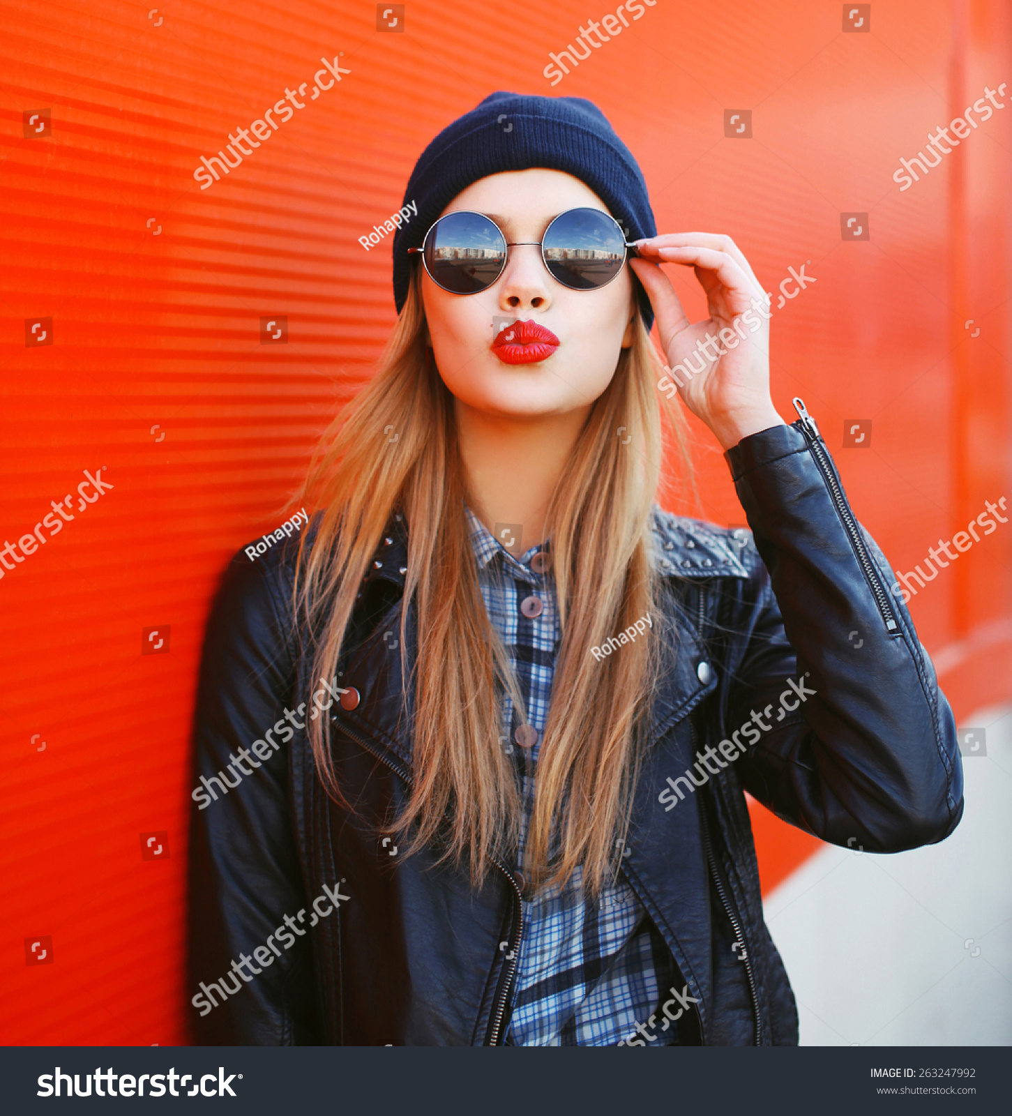 Fashionable portrait of stylish beautiful blonde young woman model blowing her lips with red lipstick sends sweet kiss in round sunglasses, black rock style leather jacket, hat on city street #263247992