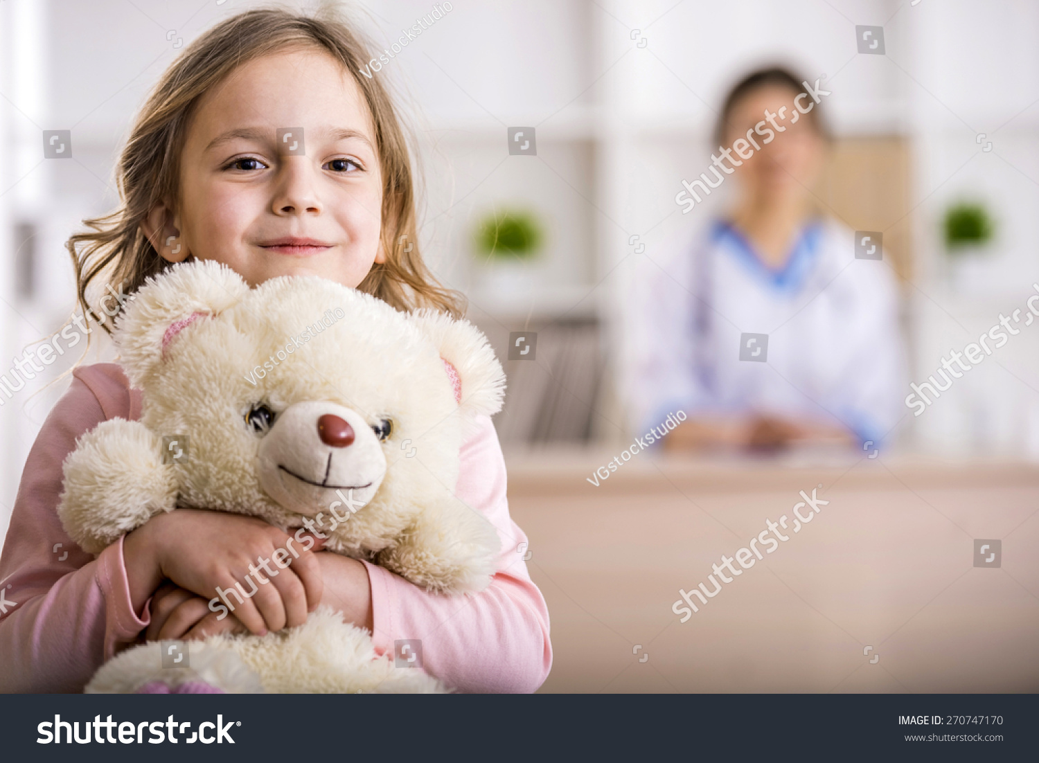 Little girl with teddy bear is looking at the camera. Female doctor on background. #270747170