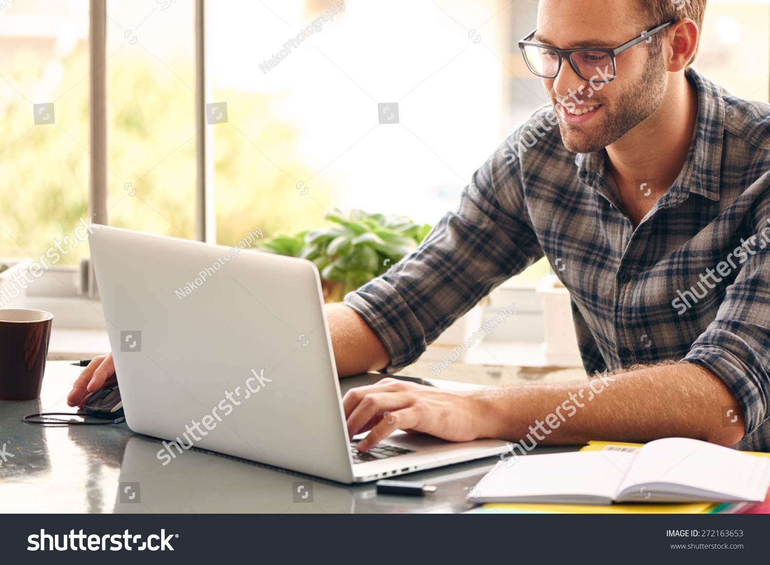 Happy young man, wearing glasses and smiling, as he works on his laptop to get all his business done early in the morning with his cup of coffee #272163653