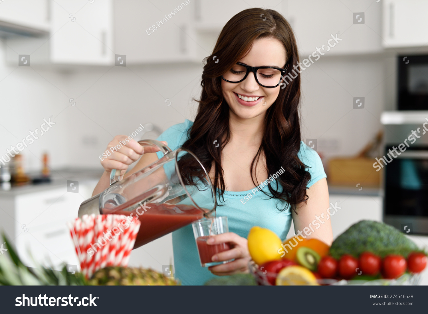 Happy healthy young woman wearing glasses pouring vegetable smoothies freshly made from assorted vegetable ingredients on her kitchen counter #274546628