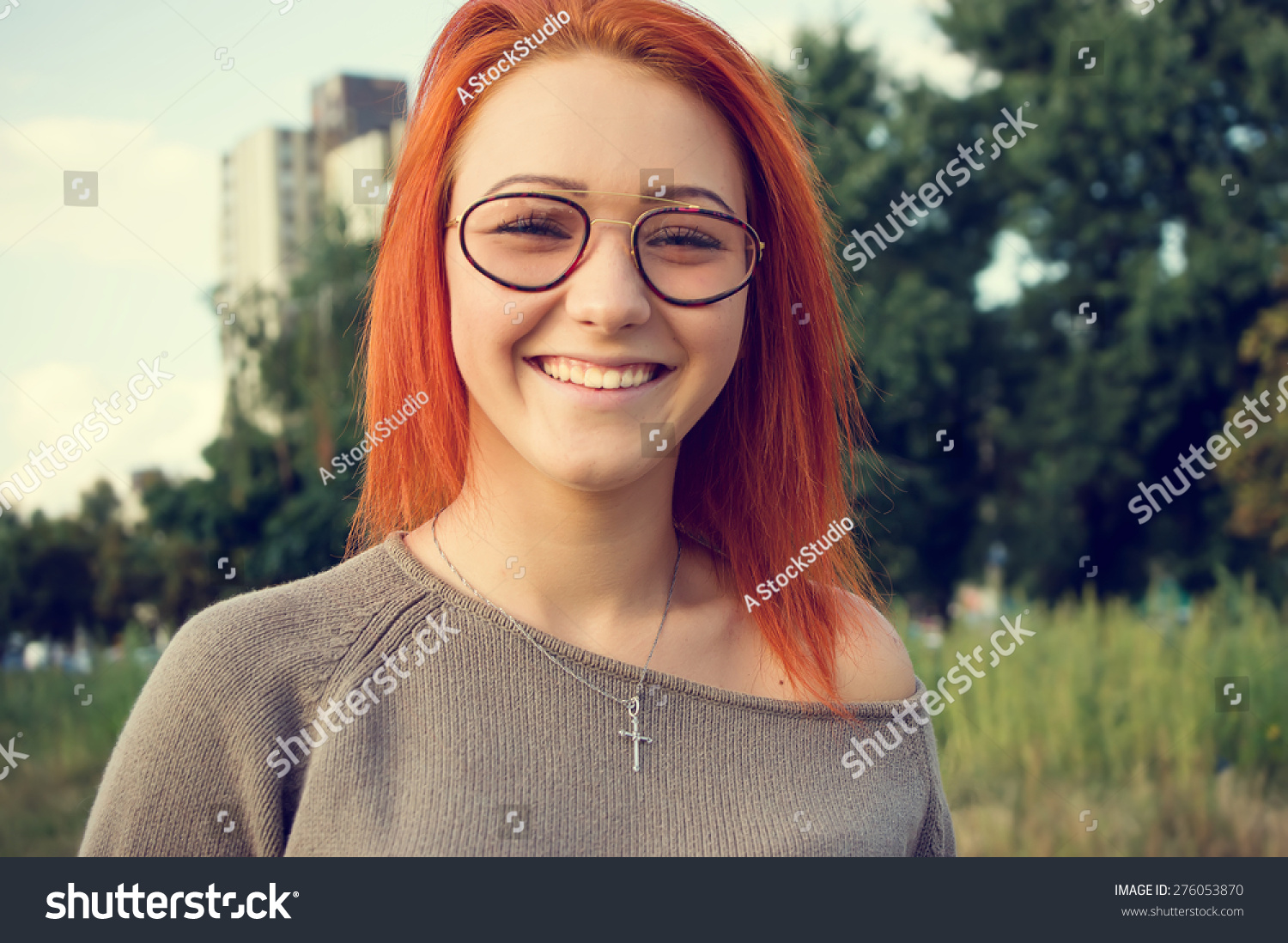 Girl with bright red hair. Woman in glasses. Beautiful happy girl outdoors. Beautiful smile on her face. Girl looks straight ahead at you. woman on the background of sky and green plants. Funny face. #276053870