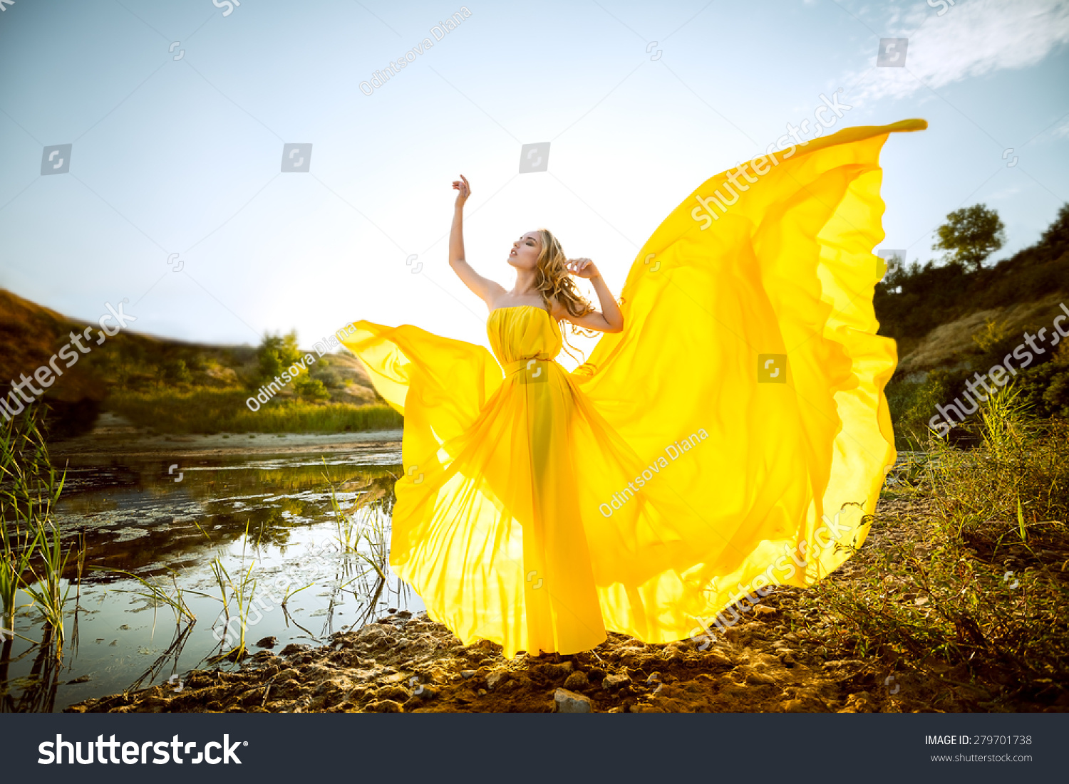 the beautiful girl with long hair in the yellow fluttering dress costs on the bank of a stream, hands raised up, having closed eyes #279701738