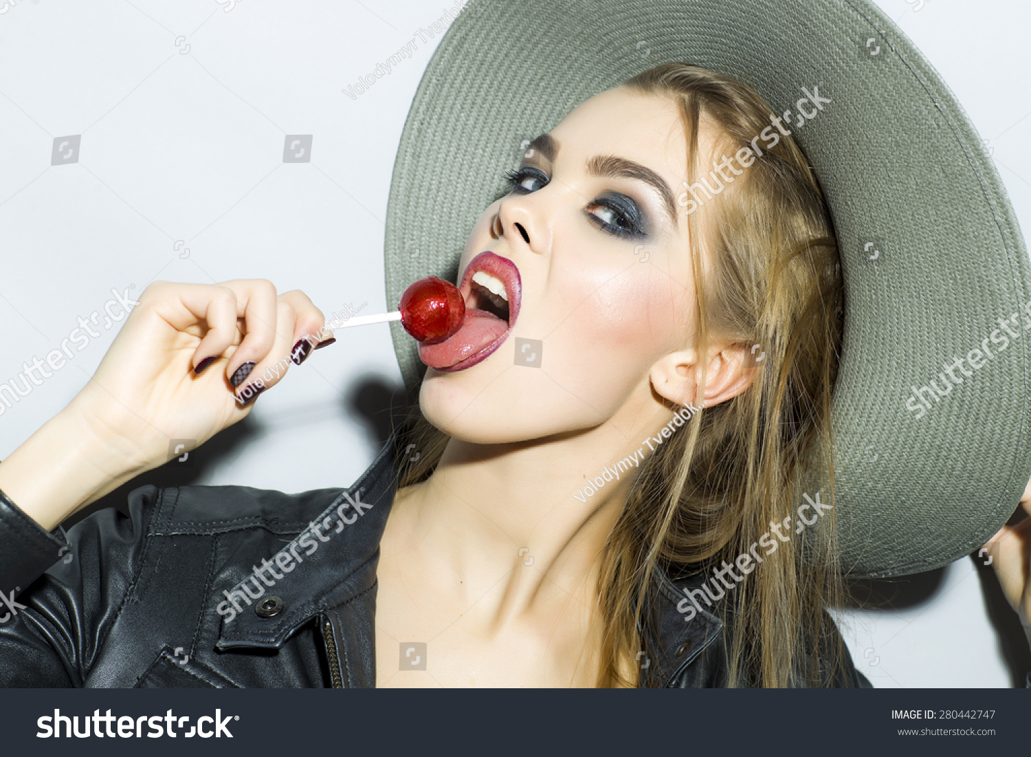 Impetuous young blonde girl portrait with bright make up in hat leather jacket and jeans looking forward holding and licking round red candy standing on gray background copyspace, horizontal picture #280442747