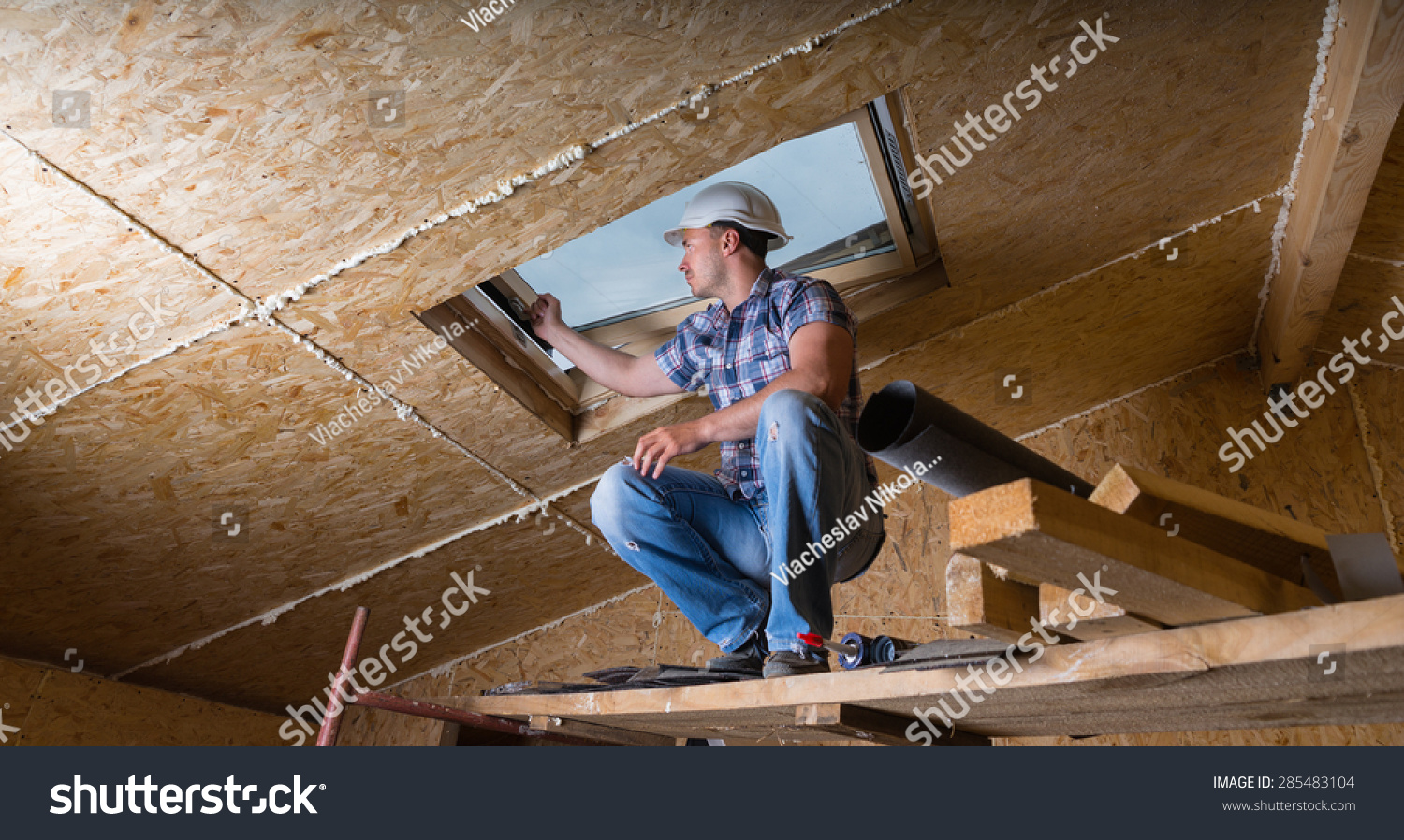 Low Angle View of Male Construction Worker Builder Crouching on Elevated Scaffolding near Ceiling and Inspecting Frame of Sky Light Window in Unfinished House with Exposed Particle Plywood Board #285483104