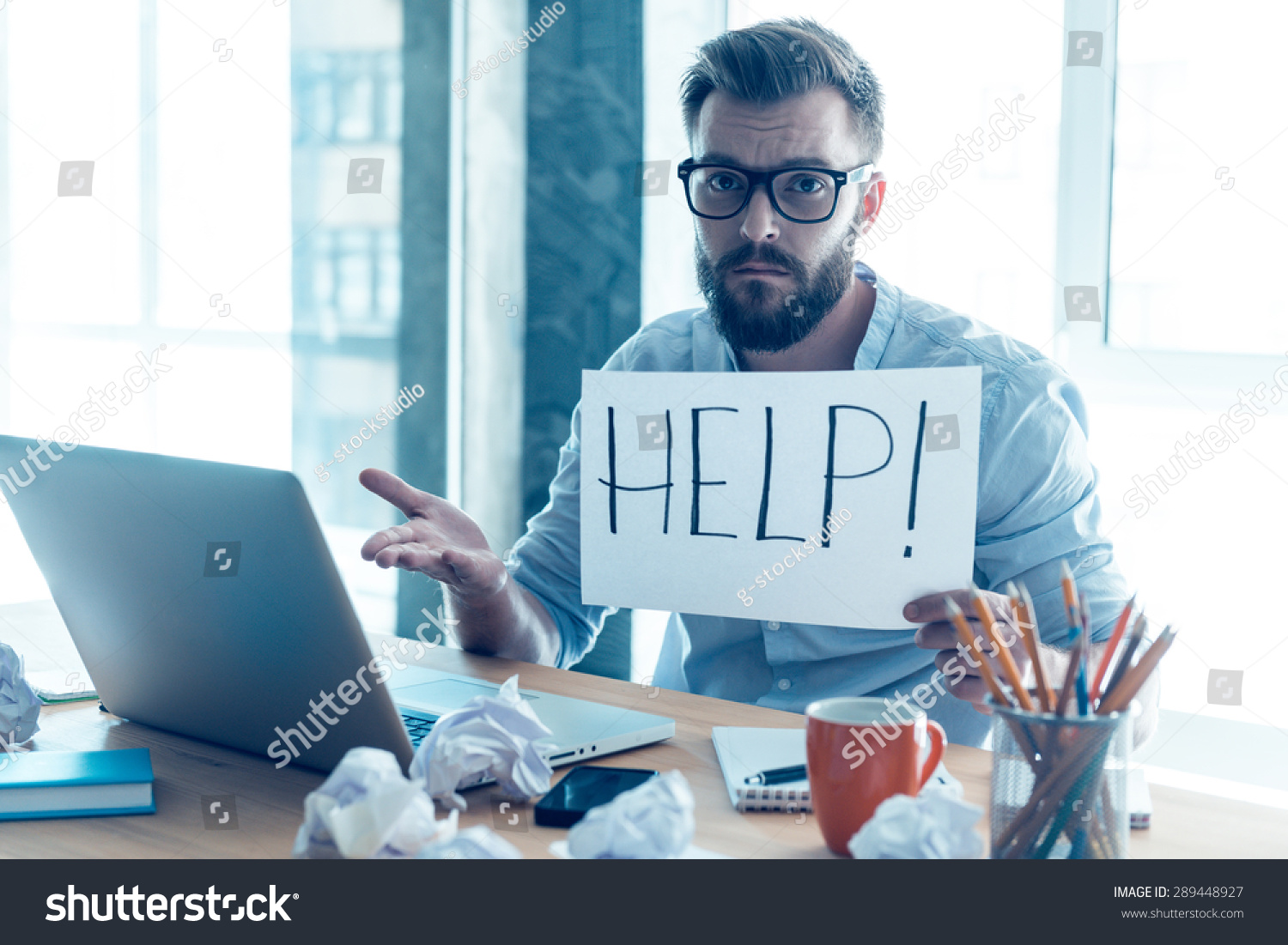 Asking for help. Frustrated young beard man holding piece of paper and asking for help while sitting at his working place in office  #289448927