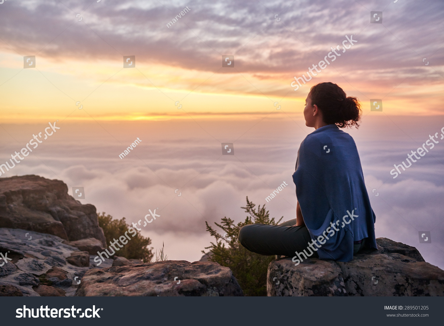 Rear view of a young woman sitting on a mountain top peacefully gazing at low-lying morning clouds and the pastel colours of a tranquil sunrise #289501205