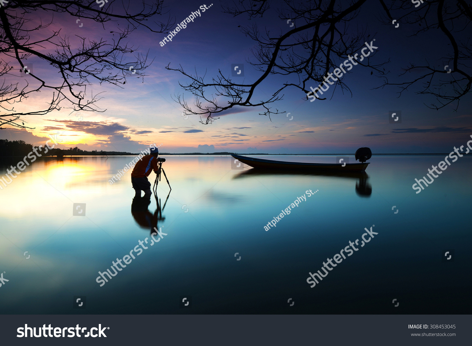 Silhouette of photographer looking to the boat near the beach when the sun goes down with branches tree border. Nature Photography #308453045