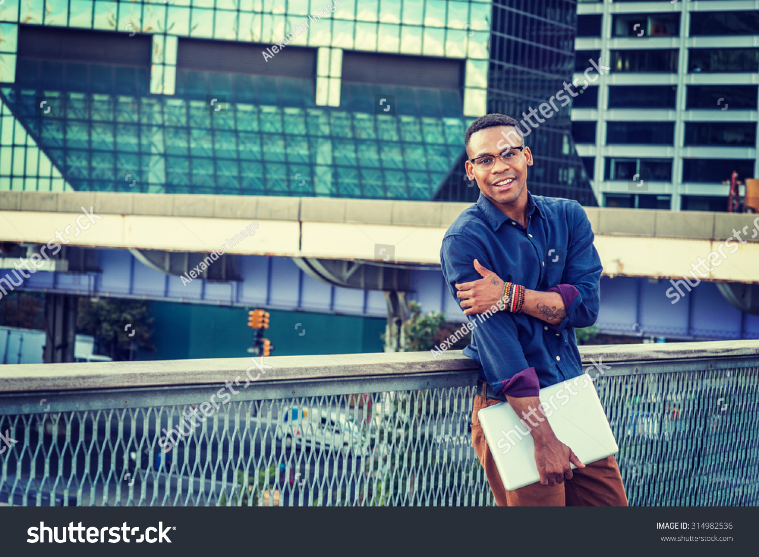 African American college student studying in New York. Wearing blue shirt, glasses, bracelets, holding laptop computer, a young black man standing in business district with high buildings, smiling.  #314982536