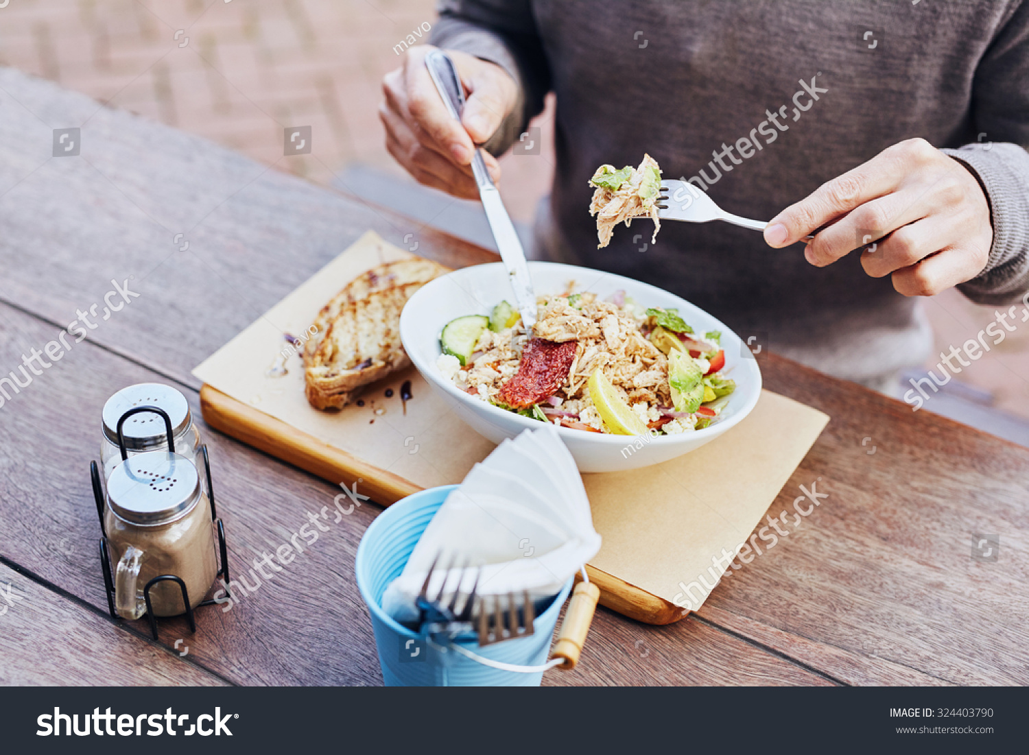 Cropped image of a man on his lunch break eating a fresh and healthy salad with chicken, avocado, sundried tomatoes and fresh sliced baguette on the side while sitting at a wooden table #324403790