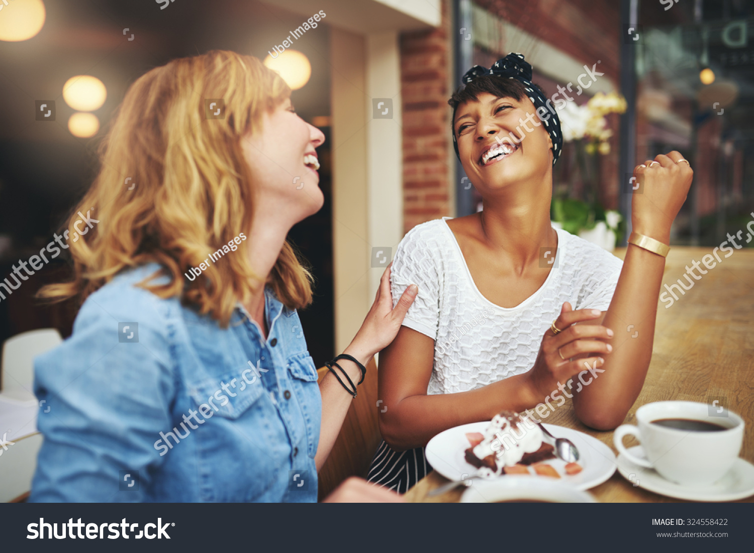 Two multiethnic young female friends enjoying coffee together in a restaurant laughing and joking while touching to display affection #324558422