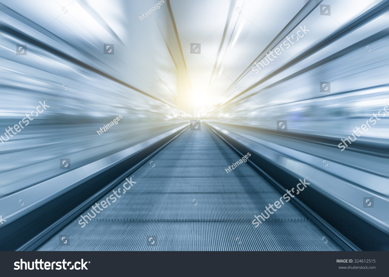 Perspective wide angle black and white view of modern light blue illuminated and spacious high-speed moving escalator with fast blurred trail of handrail in vanishing traffic motion #324612515