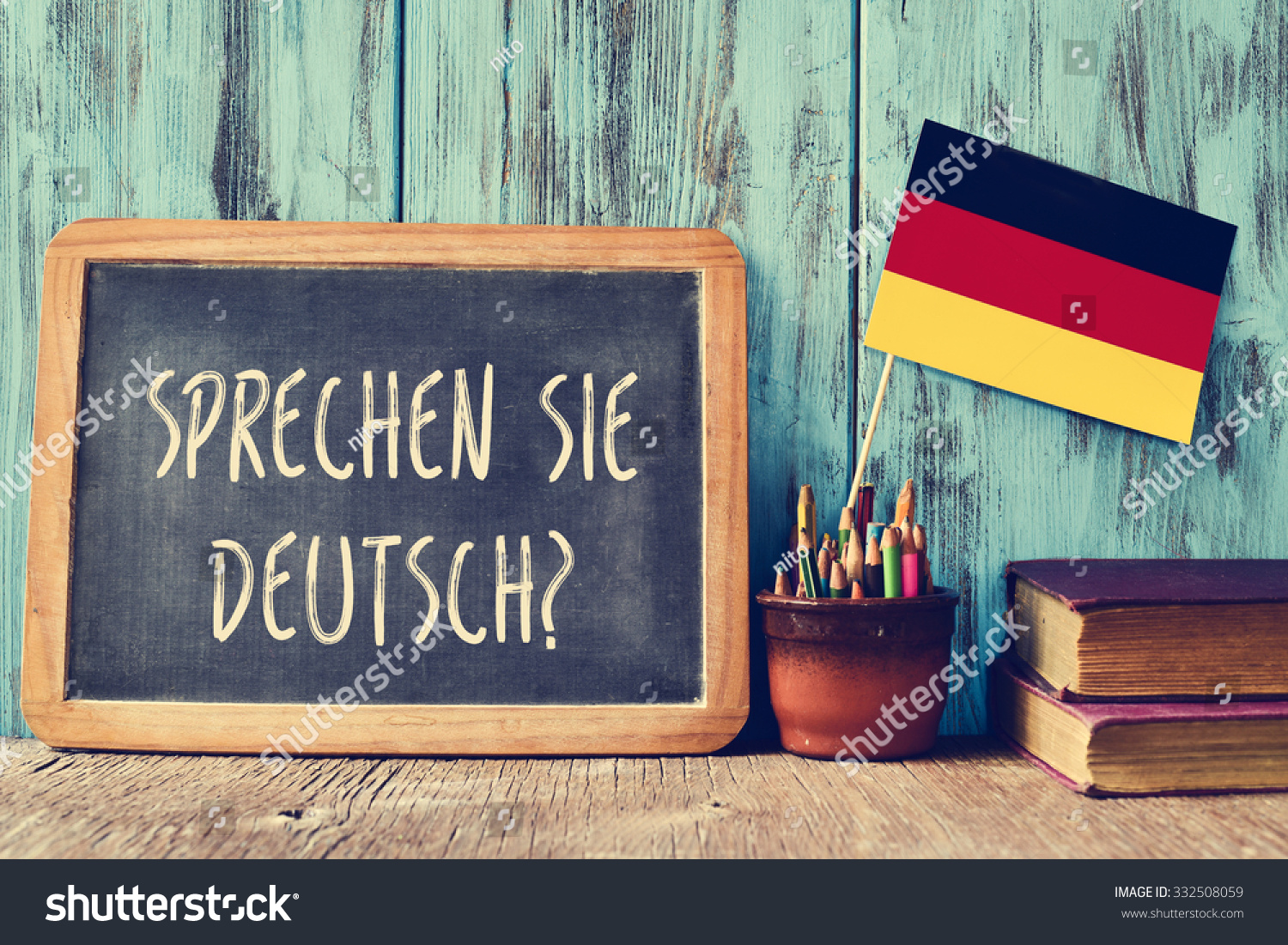 a chalkboard with the question sprechen sie deutsch? do you speak german? written in german, a pot with pencils, some books and the flag of Germany, on a wooden desk #332508059