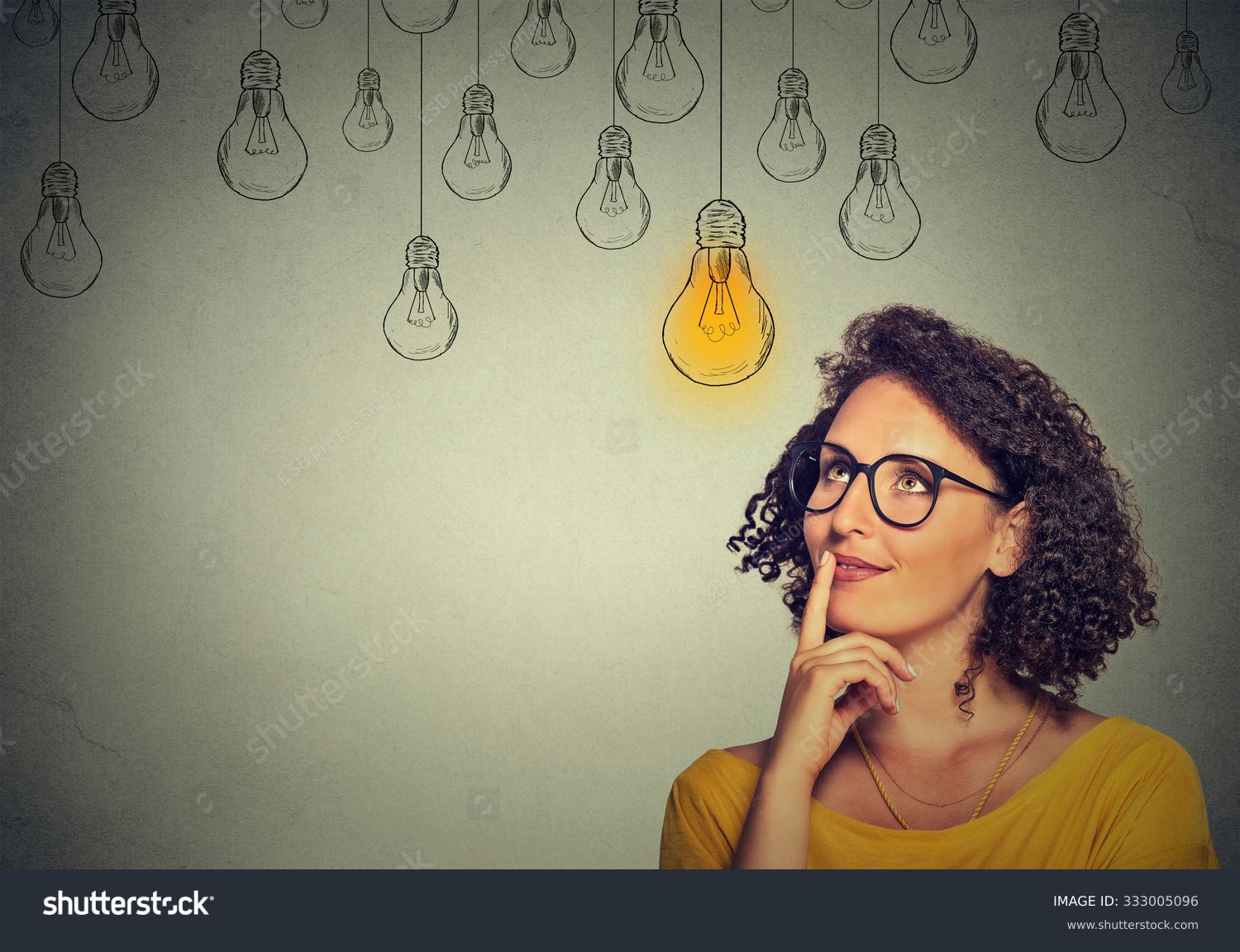 Thinking woman in glasses looking up with light idea bulb above head isolated on gray wall background #333005096