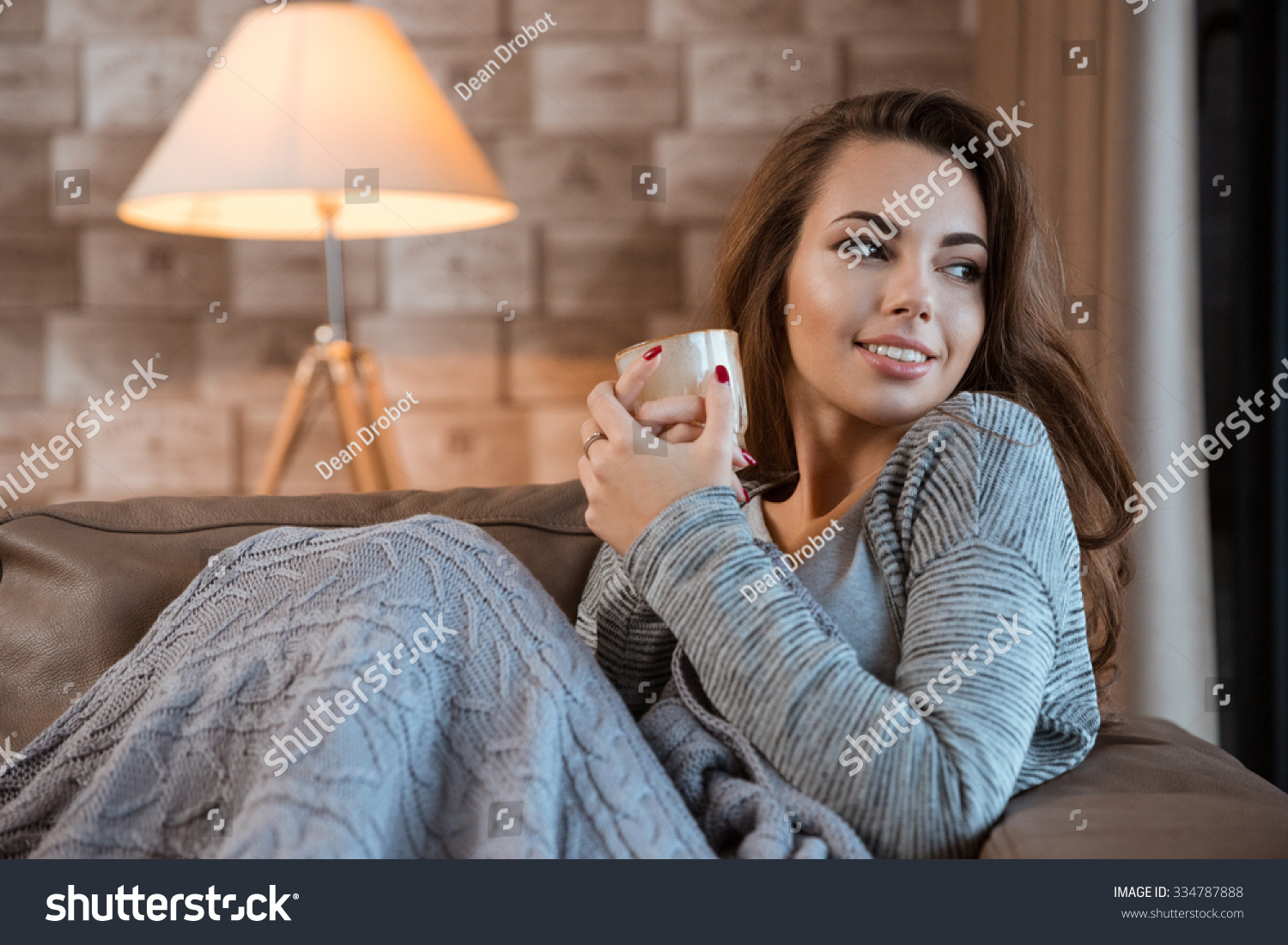 Pretty smiling girl wearing in gray clothes sitting on sofa and holding a glass #334787888