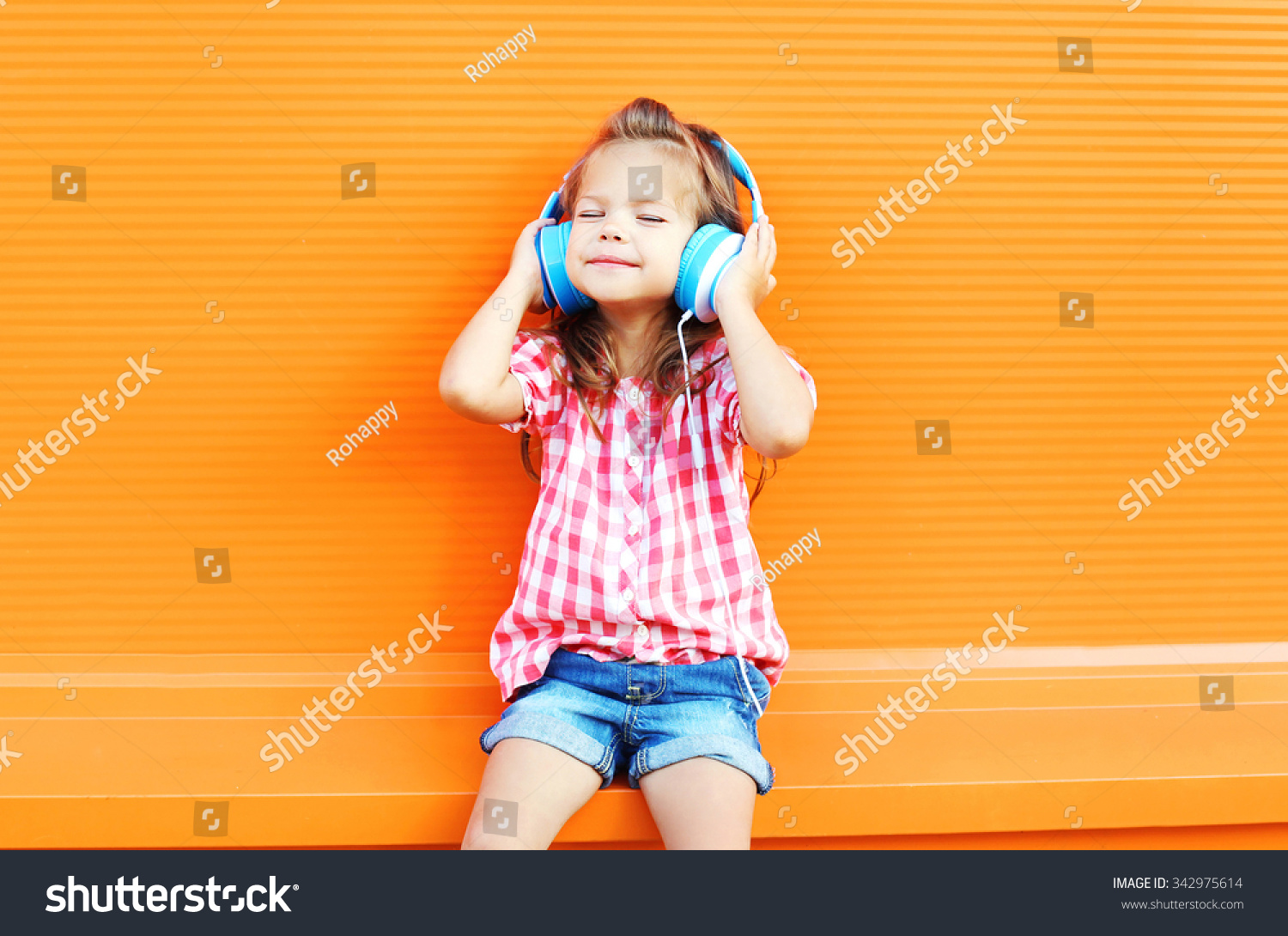 Happy smiling child enjoys listens to music in headphones over colorful orange background #342975614