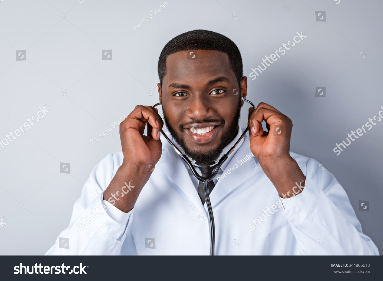 Portrait of male afro american doctor with stethoscope and lab coat. Young doctor smiling and looking at camera. Man standing on grey background #344866610