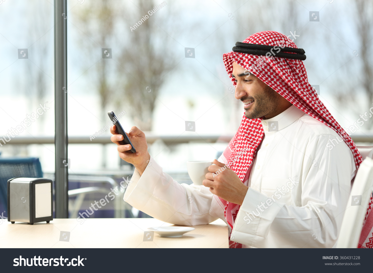 Side view of an arab man texting in a smart phone in a coffee shop with a window with a sunny day in the background #360431228