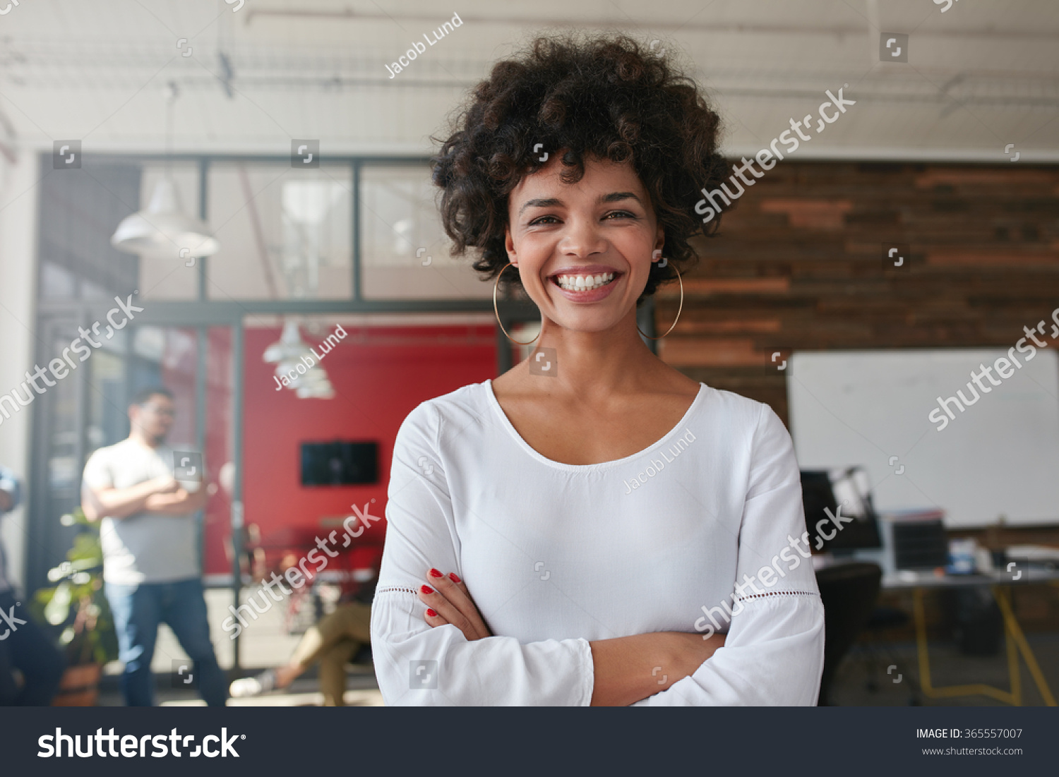 Smiling young woman standing with her arms crossed and looking at camera. She is standing in a modern office with her colleagues in the background. #365557007