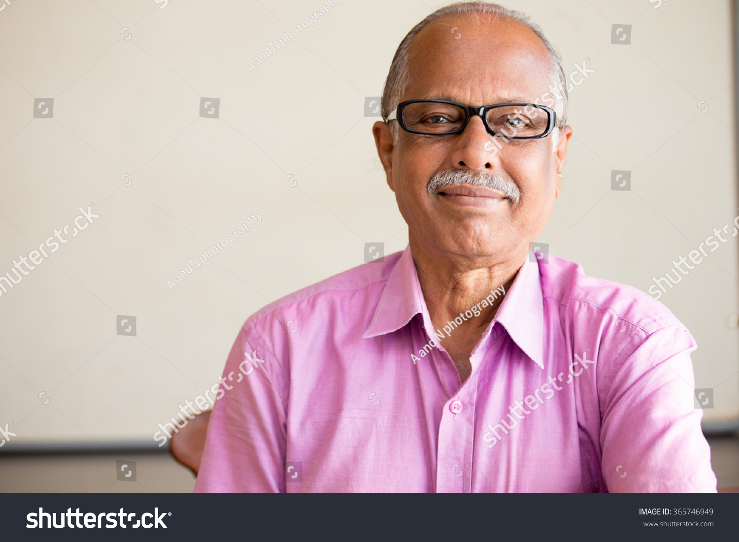 Closeup portrait, smart elderly man in pink shirt with dark eye glasses, specs, sitting down, isolated indoors white chalkboard background #365746949