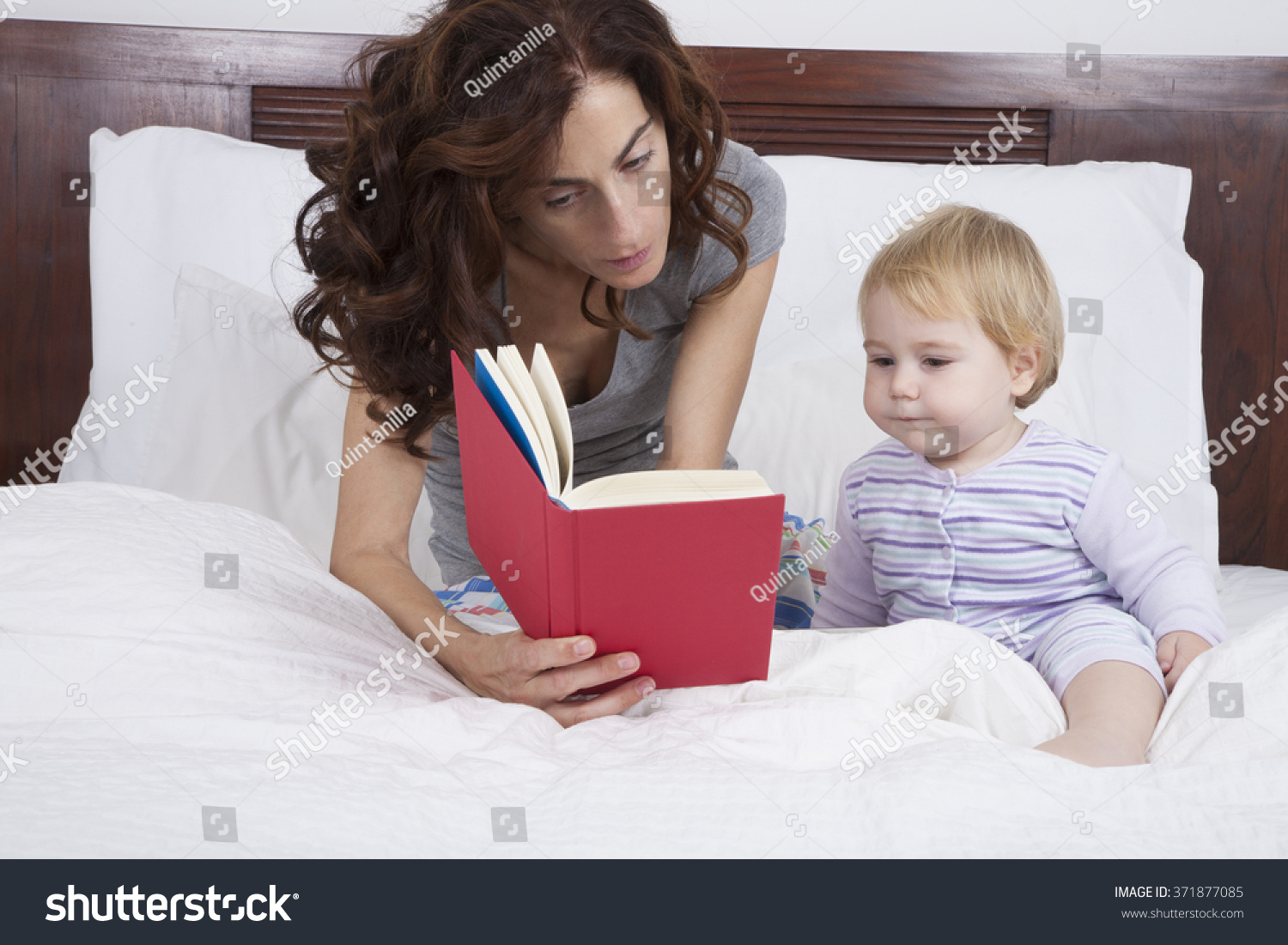 eighteen month aged blonde baby with brunette woman mother reading tale story red book in white bed #371877085
