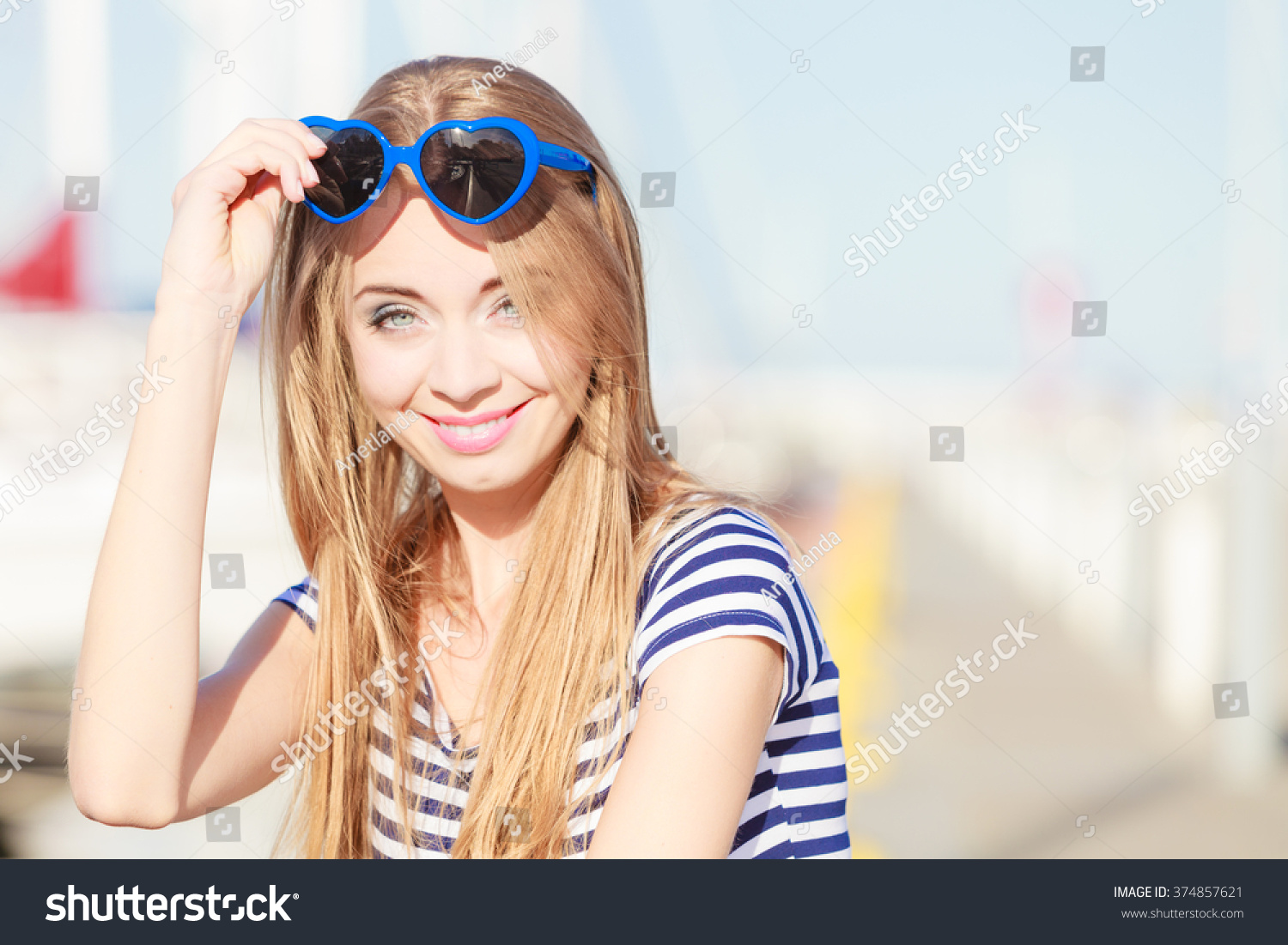 Travel tourism and people concept. Fashion blonde girl with blue heart shaped sunglasses in marina against yachts in port #374857621