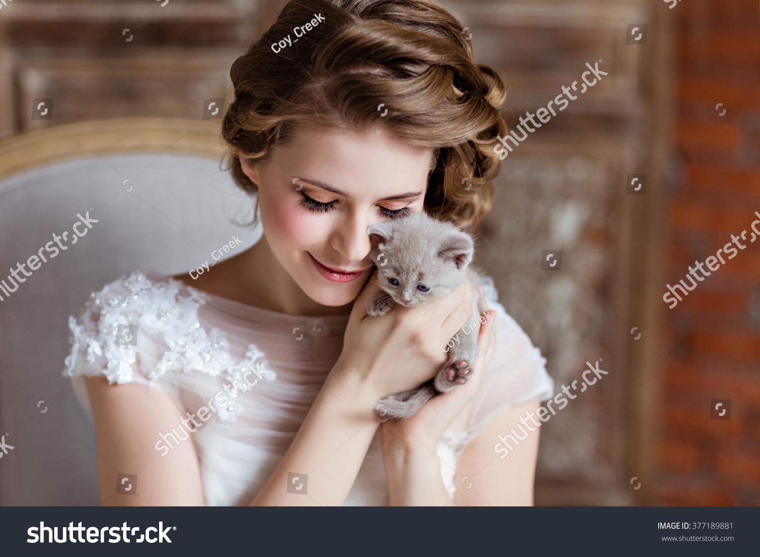 Charming and beautiful girl in white dress sitting on the couch on a background of a brick wall, smiling and holding his little kitten, close-up #377189881