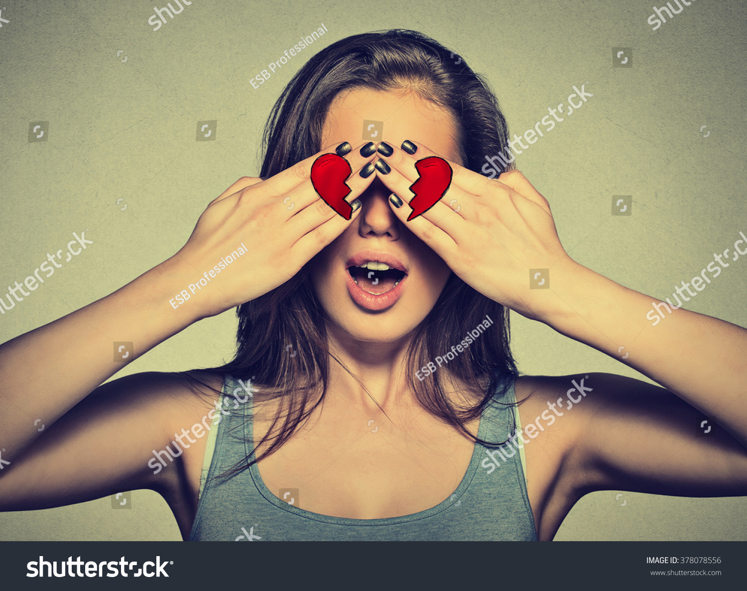Beautiful young woman eyes covered by hands with broken heart printed on them. Girl in love isolated on gray wall background    #378078556