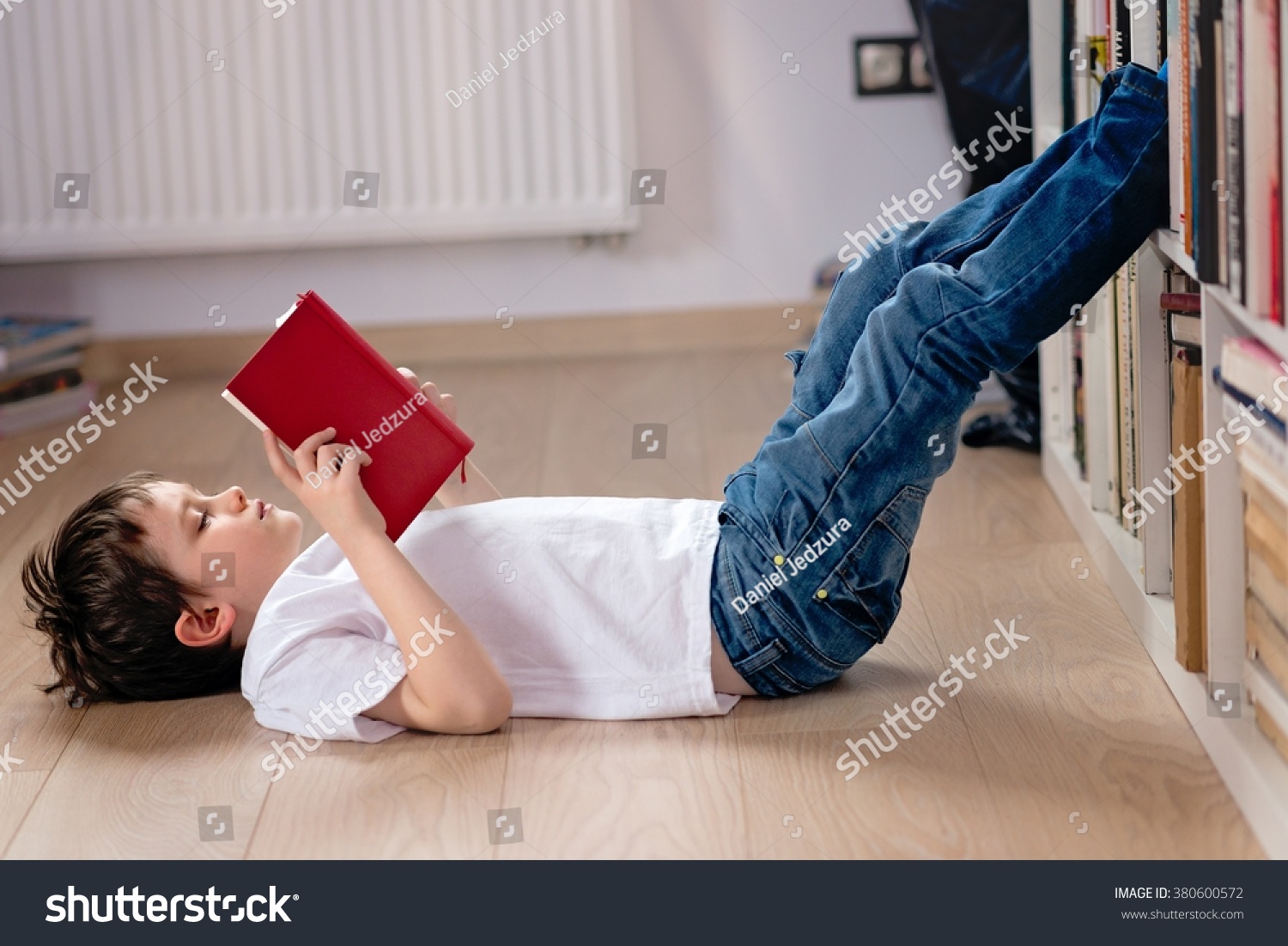 Little boy child reading a book in the library. He lies on the floor. Legs on bookshelf #380600572