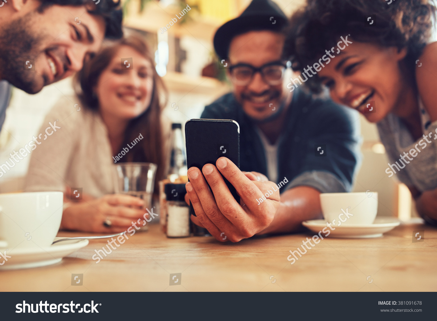 Group of friends having fun at the cafe and looking at smart phone. Man showing something to his friends sitting by, focus on mobile phone. #381091678