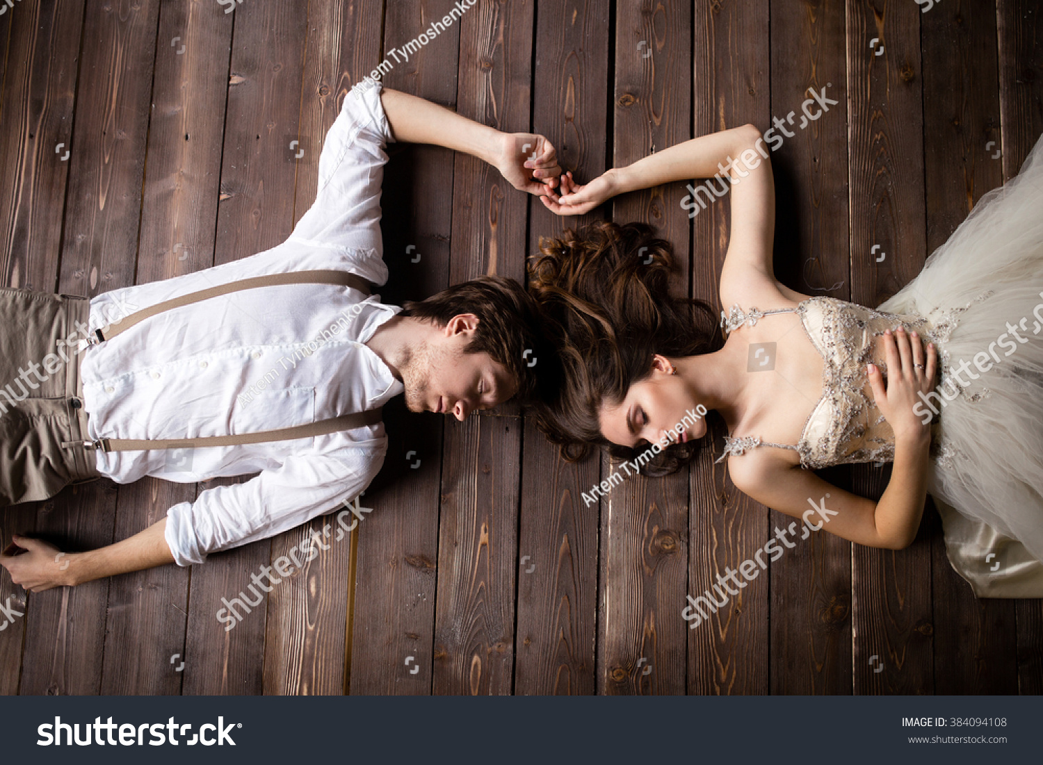 couple lying on the wooden floor and touching her hands, the bride in beige pants with suspenders and a white shirt, the bride in a beige dress #384094108
