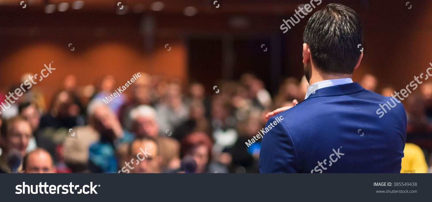 Rear view of speaker giving a talk on corporate Business Conference. Audience at the conference hall. Business and Entrepreneurship event. Panoramic composition. #385549438