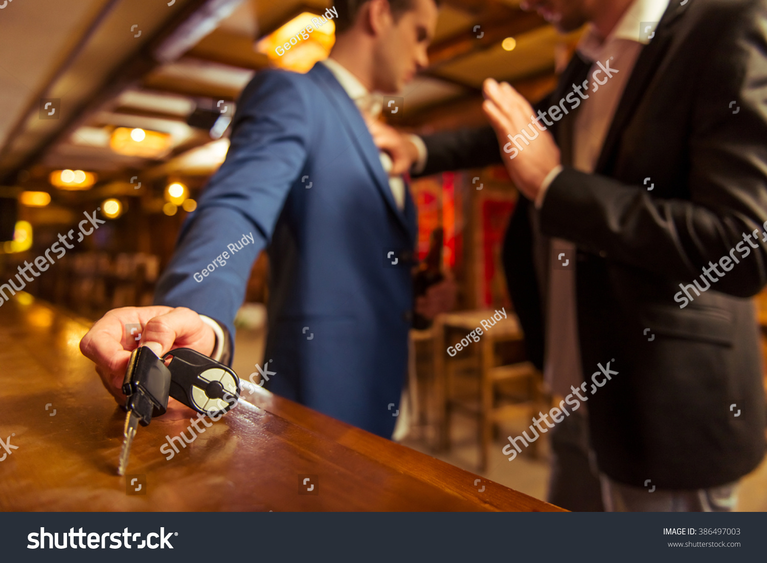 Young drunk businessman is holding a bottle of beer and reaching car keys on bar counter in pub, another man is stopping him #386497003