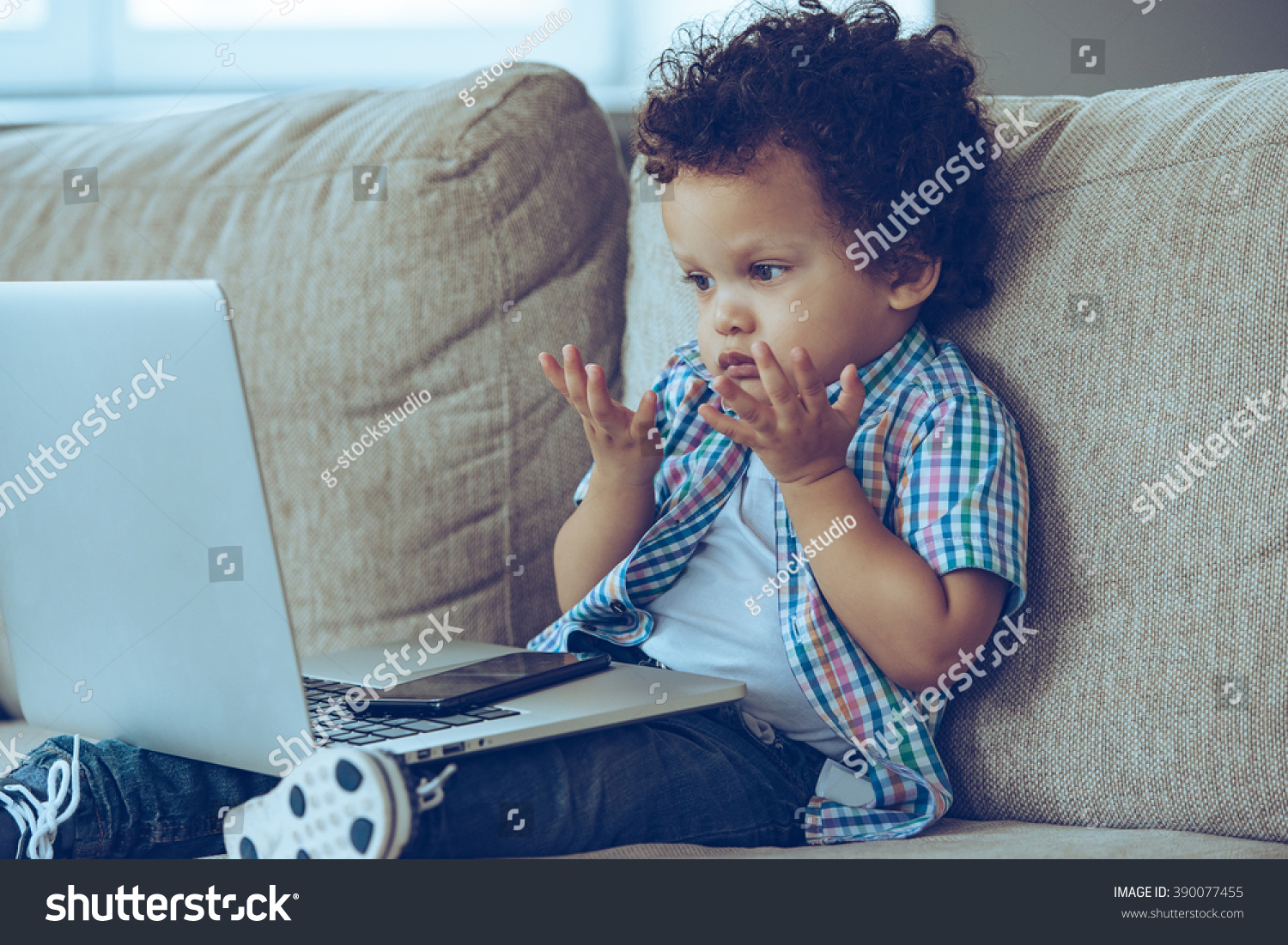 How does this thing turns on? Little African baby boy looking confused while sitting on the couch at home with laptop on his knees #390077455