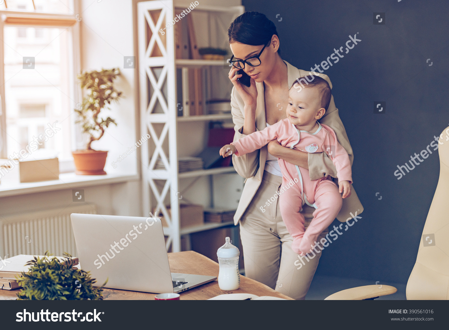 It is not easy to be a working mom! Young beautiful businesswoman talking on mobile phone and looking at laptop while standing with her baby girl at her working place #390561016