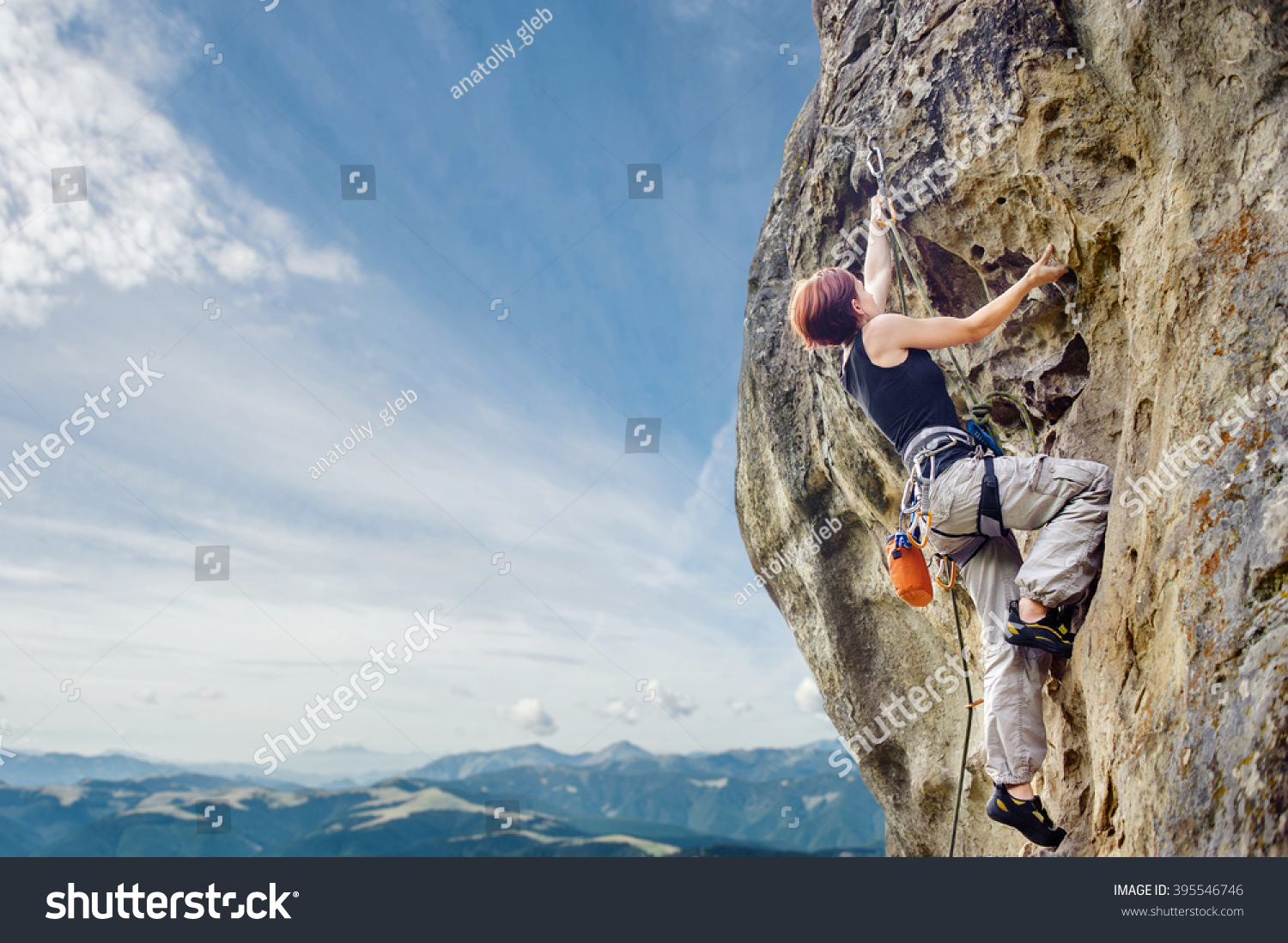 Rear view of young female climber climbing with rope and carbines, looking for next grip on a big rocky wall against blue sky and mountains. Summer time. Climbing equipment #395546746