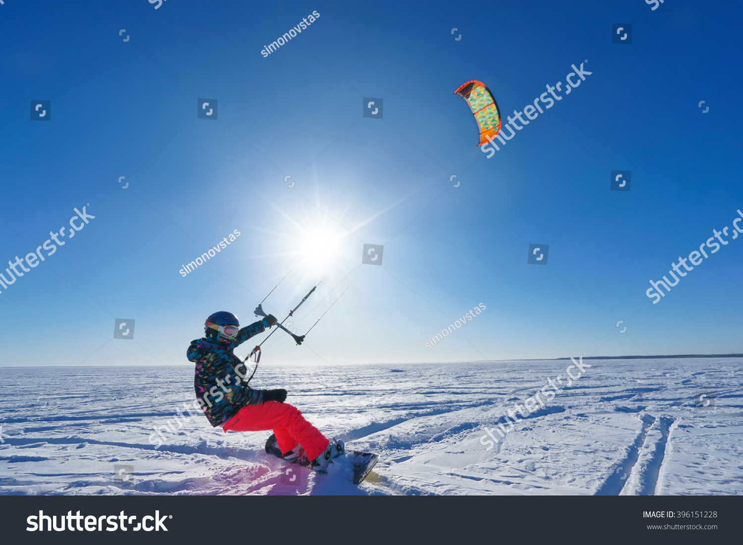 Athlete in bright clothes riding in the snow on a snowboard and kite controls. Deep blue sky and sun in the background #396151228
