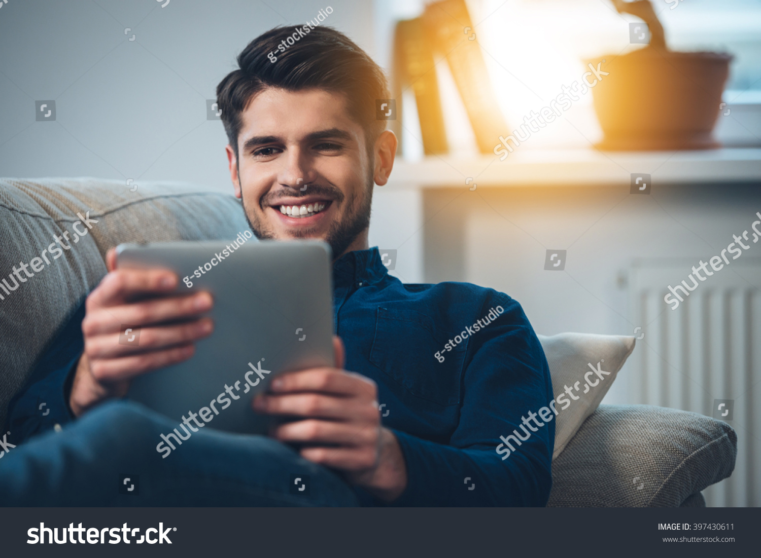 Spending great time at home. Close-up of handsome young man using his digital tablet with smile while lying down on the couch at home #397430611