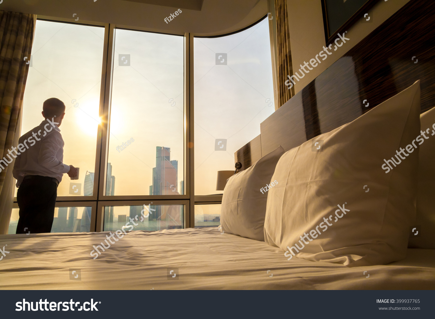 Bed maid-up with white pillows and bed sheets in cozy room. Young businessman with cup of coffee standing at window looking at city scenery on the background. Focus on cushion. Motivation concept #399937765