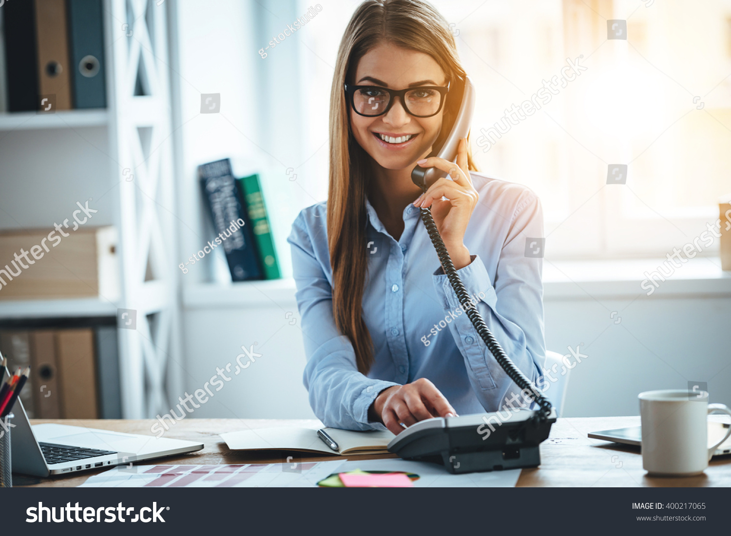 I will connect you in one second! Cheerful young beautiful woman in glasses talking on the phone and looking at camera with smile while sitting at her working place #400217065