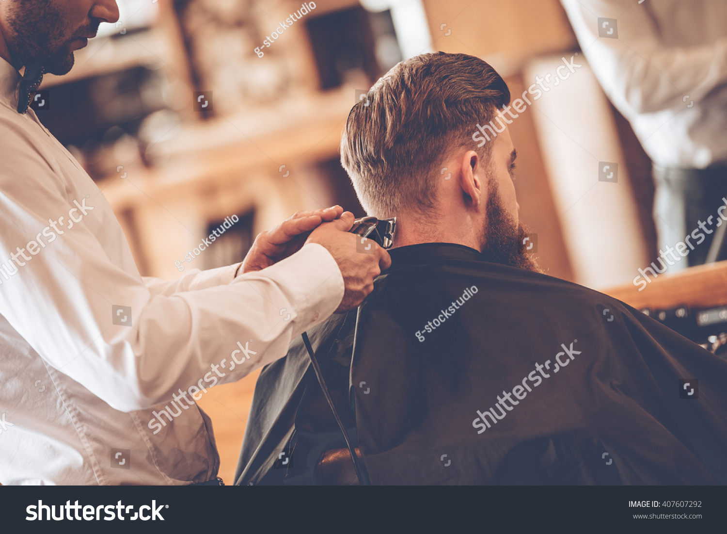 Perfect  trim. Rear view close-up of young bearded man getting haircut by hairdresser with electric razor while sitting in chair at barbershop #407607292