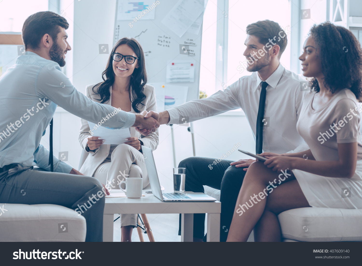 Welcome on board! Two handsome men shaking hands with smile while sitting on the couch at office with their coworkers #407609140