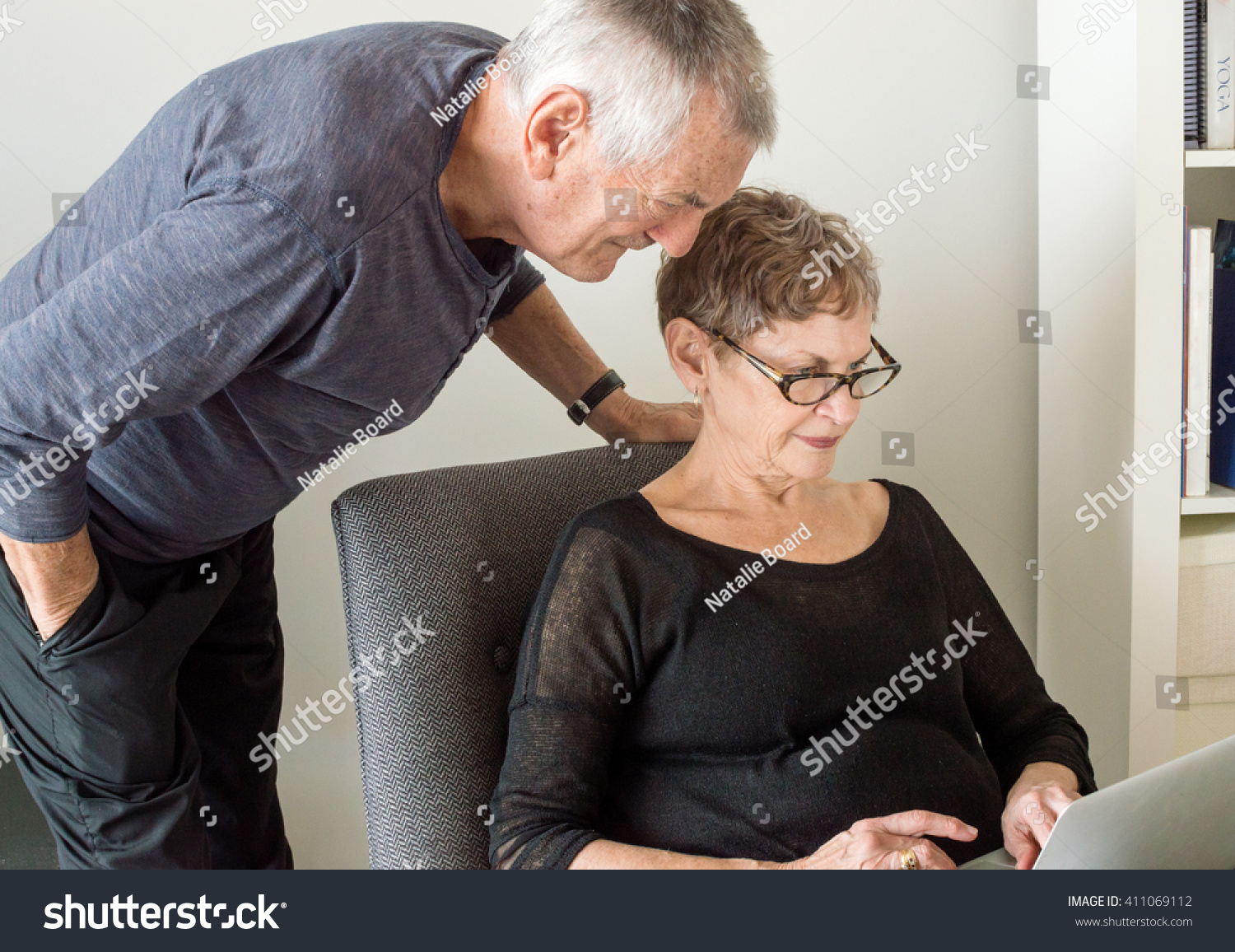 Older woman in black clothing seated using a computer with older man in dark clothing looking over her shoulder #411069112